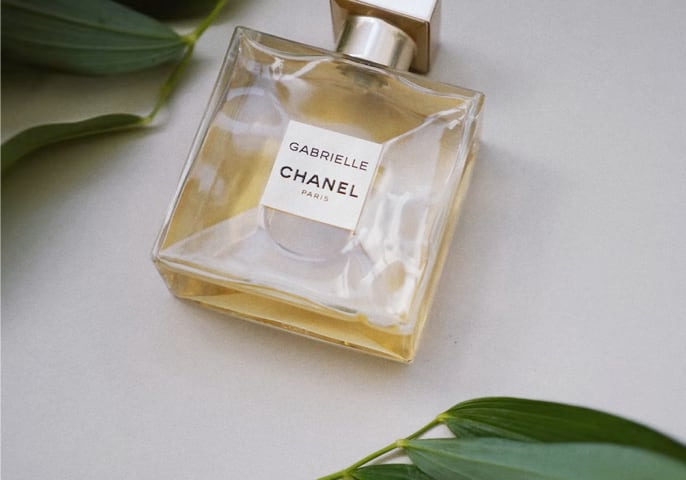 A bottle of Gabrielle Essence Eau De Parfum lies against a plain background, with leaves above and below.